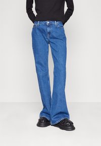 Blue denim flared jeans with a mid-rise waist, five pockets, and a single front button closure, paired with black textured slip-on shoes.