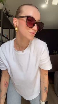 Pessoa com cabelo puxado para trás usa grandes óculos de sol castanhos, brincos de argola dourados, uma t-shirt branca, calças às riscas, e tem tatuagens em ambos os braços.