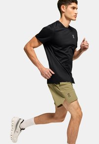 Schwarzes Sport-T-Shirt und olivgrüne Shorts, Läufer in Aktion. Strukturierter Stoff, Logodetail auf den Shorts, weiße Socken und Sneakers.