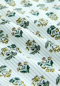 Tissu blanc côtelé avec de petits motifs floraux jaunes et verts dispersés sur toute la surface, présenté en plis doux.