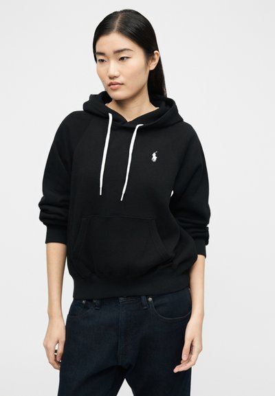 Νεαρή γυναίκα που φοράει μαύρη φούτερ με λευκά κορδόνια και σκούρο τζιν, όρθια μπροστά σε ένα απλό ανοιχτό φόντο, κοιτάζοντας προς τα κάτω και πλάγια.