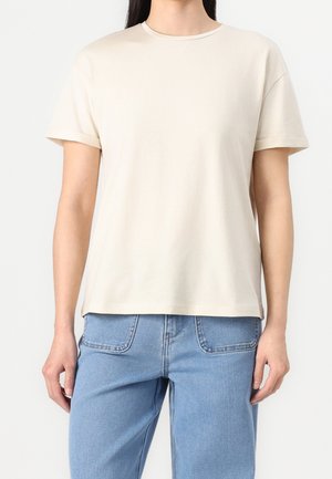 Osoba ubrana w gładki beżowy t-shirt z krótkim rękawem w zestawieniu z jasnoniebieskimi jeansami, stojąca na białym tle.