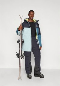 Skijas in lichtblauw en donkerblauw met een capuchon, voorzien van meerdere zakken en een rits. Zwarte skibroek en laarzen. Ski's in de hand gehouden.