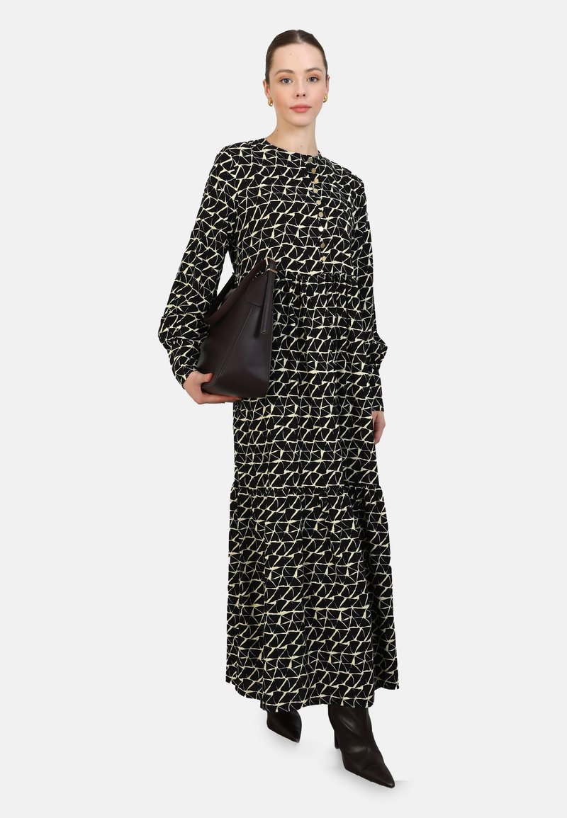 Schwarzes und cremefarbenes, gemustertes Maxikleid mit langen Ärmeln und gerafftem Bund, kombiniert mit einer dunkelbraunen Handtasche und spitzen, schwarzen Stiefeln.