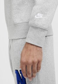 Šedá bavlněná mikina s žebrovanými manžetami a bílým logo Nike. Detailní záběr ukazuje ruku a modrou vyšívanou nášivku na šedých kalhotách.