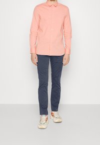 Hellrosa langärmeliges Hemd mit Kragen, kombiniert mit dunkelblauen Hosen und mehrfarbigen Turnschuhen mit Wellenmustern und klobigen Sohlen.