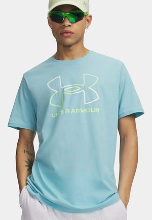 Junger Mann trägt ein hellblaues Under Armour T-Shirt, eine hellfarbige Kappe, neongrüne Sonnenbrille und weiße Shorts, mit sichtbaren Tätowierungen am linken Arm.