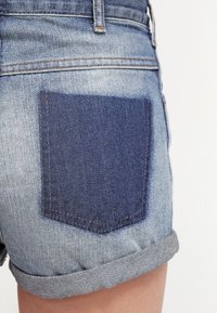 Džínové šortky s světle modrým praním a tmavě modrou nášivkou na kapsu. Otočený lem a viditelné švy na pase a okrajích.