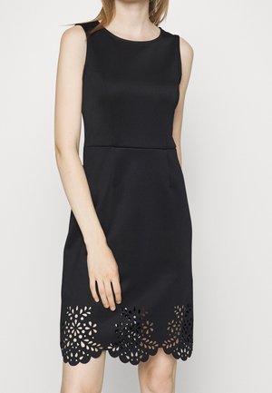 Ärmelloses schwarzes knielanges Kleid mit rundem Ausschnitt und floral ausgeschnittenem Saum, getragen von einer stehenden Person vor einem einfarbigen Hintergrund.