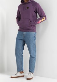 Fialová mikina s kapucí a přední kapsou, logo na hrudi a grafický vzor na rukávech. Spárována se světle modrými džínami a bílými teniskami.