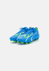 Botas de fútbol azules con una superficie texturizada, acentos en verde neón, detalles en blanco y tacos moldeados en la suela para una mejor tracción.