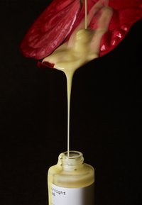 Flaske med bred hals, klart glass, hvit etikett, viskøs gul væske som drypper fra en rød, kronbladlignende struktur på flasken.