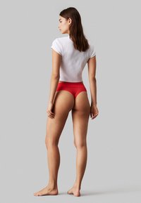 Röd underkläder med en texturerad midja, kombinerat med en vit t-shirt med kort ärm. Modellen står barfota och visar upp en åtsittande design.