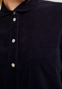 Camicia in velluto a coste blu navy con colletto, dotata di bottoni a pressione argentati e taschino sul lato sinistro del petto. Texture a coste e design strutturato.