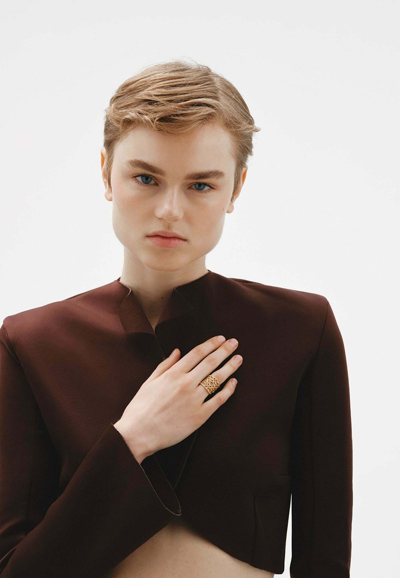 Chaqueta marrón corta con un cuello alto, combinada con un anillo dorado intrincado en la mano. Textura suave, diseño minimalista.