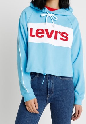 Ljusblå kort hoodie med en vit remsa med röd "Levi's"-logotyp, dragsko i huvan och vita sidostycken. Kombinerad med blå jeans.