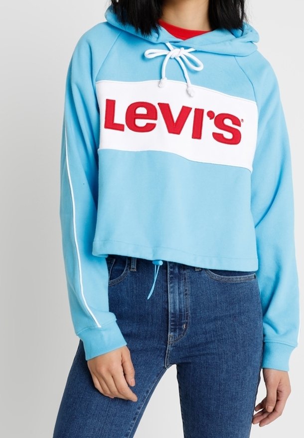 Felpa cropped azzurro chiaro con una banda bianca e il logo rosso "Levi's", cappuccio con coulisse e dettagli bianchi a strisce laterali. Abbinata a jeans blu.
