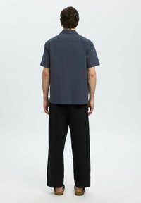 Mann som står med ryggen til, iført en teksturert kortermet mørkegrå skjorte, løse svarte bukser og brune sandaler på en hvit bakgrunn.