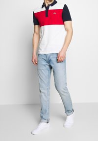 Polo pour homme avec base blanche ornée de rayures horizontales rouges et bleu marine, manches courtes et patte de boutonnage à trois boutons. Porté avec un jean bleu clair.