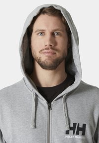 Sudadera con cremallera en gris, fabricada en una mezcla de algodón, que cuenta con un bolsillo frontal, capucha ajustable con cordón y un logo negro en el lado izquierdo del pecho.