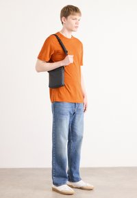 Melnas, taisnstūra formas pleca soma, izgatavota no auduma ar regulējamu siksnu, kuru tur persona oranžā krekla un zilos džinsos. Vienkāršs dizains.