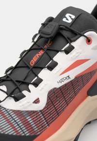 Trail running shoe with a black and red textile upper, white accents, distinctive checkered pattern, elastic laces, and a reinforced sole.