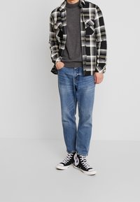 Mann in einem schwarz-weißen karierten Hemd, grauem Rollkragenpullover, blauen Jeans und schwarzen High-Top-Sneakern, der vor einem schlichten Hintergrund steht.