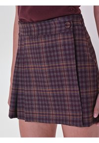 Jupe écossaise prune avec une fermeture à boutons, des plis et des lignes orange contrastantes sur un tissu texturé. Longueur courte et taille ajustée.