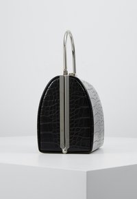 Pieni musta käsilaukku krokotiilinkuviolla, hopeinen metallinen lukko ja pyöreä kahva, valkoisen jalustan päällä yksinkertaisen taustan edessä.