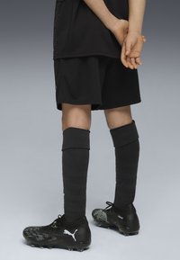 Kind steht mit hinter dem Rücken verschränkten Händen da, trägt schwarze Sportshorts, hohe schwarze Socken und schwarze Puma-Fußballschuhe.