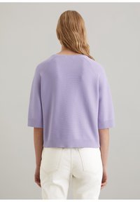 Lavendelfarbenes kurzärmliges Strickoberteil mit Rundhalsausschnitt und gerippten Bündchen, kombiniert mit hellen Hosen. Glatte Textur und entspannter Schnitt.