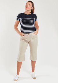 Niebiesko-białe w paski t-shirt z krótkim rękawem zestawiony z beżowymi spodniami 3/4, noszony z białymi trampkami. Model stoi z rękami w kieszeniach.