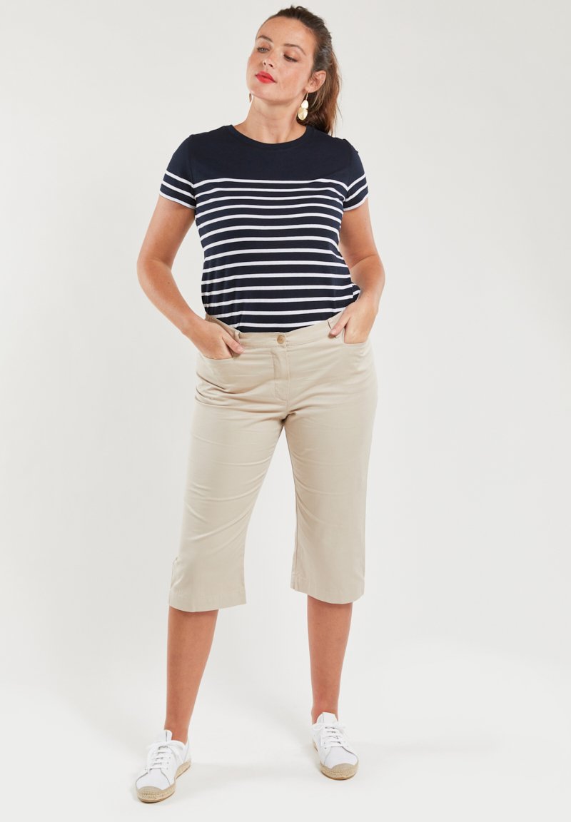 Niebiesko-białe w paski t-shirt z krótkim rękawem zestawiony z beżowymi spodniami 3/4, noszony z białymi trampkami. Model stoi z rękami w kieszeniach.