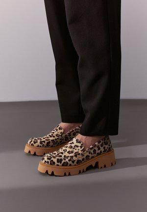 Buty wsuwane w panterkę z teksturowanej tkaniny, z grubą gumową podeszwą i głębokimi rowkami dla lepszej przyczepności. Noszone z czarnymi spodniami.