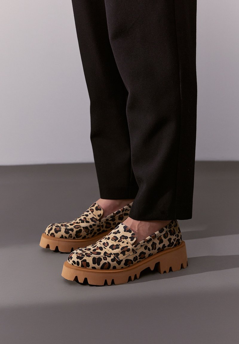 Lederimitat-Slipper mit Leopardenmuster und einem strukturierten Obermaterial, robuster Gummisohle und tiefen Rillen für besseren Halt. Getragen mit schwarzen Hosen.