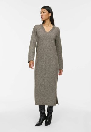 Mujer con un vestido de punto largo gris con mangas largas y escote en V, combinado con botas negras de punta afilada, que está de pie frente a un fondo blanco.