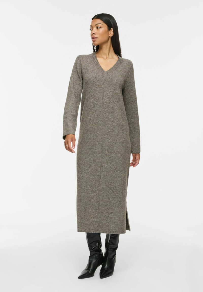 Femme portant une longue robe en maille grise à manches longues et col en V, associée à des bottes noires à bout pointu, debout devant un fond blanc.