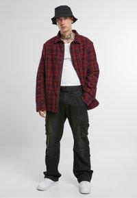 Červeno-čierna károvaná flanelová košeľa vrstvená cez biele tielko, skombinovaná s čiernymi cargo nohavicami. Čierny bucket hat a biele tenisky dotvárajú outfit.