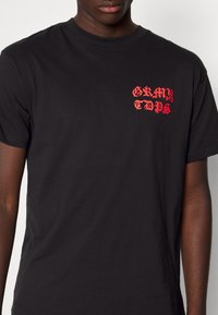 Černé bavlněné tričko s červeným grafickým textem "GRMY TDPS" na levém prsu. Krátké rukávy a kulatý výstřih. Hladká textura.
