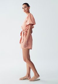 Peachi värvi lühike kleit, mis on valmistatud kergest kangast, omab puhvärve, vööd ja kahte esist taskut. Kandmine koos rihmaliste sandaalidega.