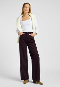 Weißes Langarmoberteil, das mit einer beigen Strickweste kombiniert ist, dazu hochgeschnittene, weit geschnittene burgunderfarbene Hose und beige Ankle Boots mit Blockabsatz.