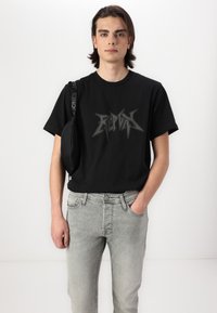 Svart t-skjorte med kort erme og fet grå grafisk tekst; kombinert med lysegrå denim jeans. En liten svart veske henger fra skulderremmen.