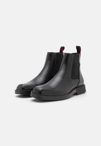 Bottes Chelsea en cuir noir avec panneaux élastiques et languettes de tirage. Bout arrondi, talon plat, et finition lisse avec des accents rouges contrastants.