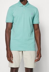 Svetle tyrkysové polo tričko s klasickým límečkem, krátkými rukávy a malým purpurovým logem na hrudi. Nosené s světle béžovými šortkami.