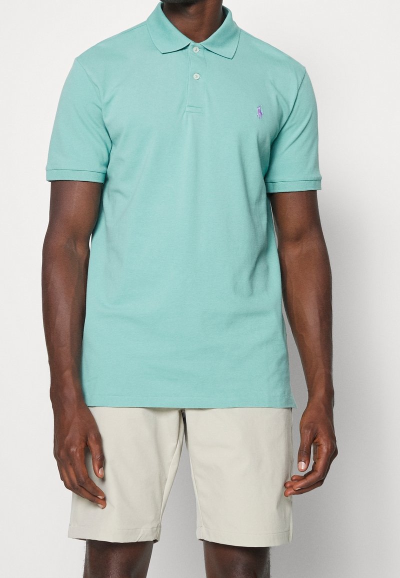Svetle tyrkysové polo tričko s klasickým límečkem, krátkými rukávy a malým purpurovým logem na hrudi. Nosené s světle béžovými šortkami.