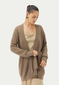 Brun stickad cardigan med diamantmönster, öppen front, långa ärmar och ett knytbälte med rektangulär spänne. Bärs över en beige topp.