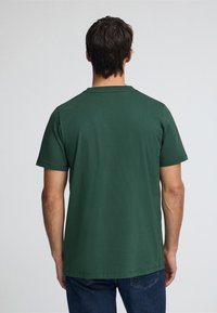 Grön bomulls-T-shirt med rund halsringning och korta ärmar, mjuk textur och rak fåll, kombinerad med blå jeans, sedd bakifrån.