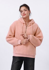 Roza flis dukserica s patentnim zatvaračem, oversized kroja i kapuljačom s vezicom. Rebrasti manžeti i rub, mekana tekstura, suptilni sjaj.