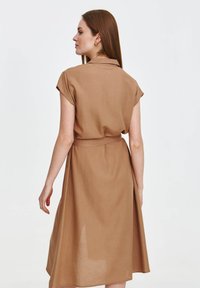 Krótkosleeve'owa, brązowa sukienka z lnu z kołnierzykiem, paskiem w talii i luźnym krojem, sięgająca do połowy łydki, o gładkiej fakturze i minimalistycznym wzornictwie.