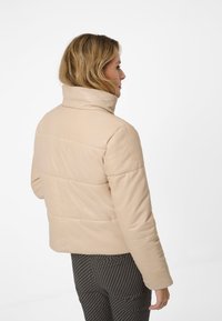 Beige vadderad jacka med hög krage; slät textur; horisontell stickning; kort silhuett; bärs med mönstrade svarta leggings.